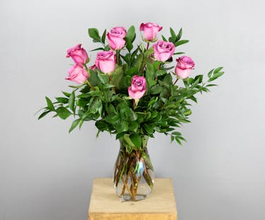 This arrangement features a dozen lavender roses elegantly arranged in a sleek clear glass vase, accompanied by assorted greenery.