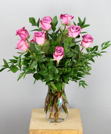 This arrangement features a dozen lavender roses elegantly arranged in a sleek clear glass vase, accompanied by assorted greenery.